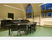 Innenraum eines Lesesaals oder Studienraums mit hohen Decken und großen, bogenförmigen Fenstern. Im Vordergrund stehen schwarze Tische und gepolsterte Stühle, im Hintergrund ein Pult mit Büchern, Arbeitsplätzen und einer Garderobenständer.