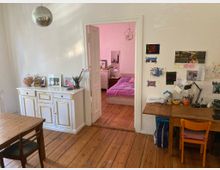 Wohnzimmer mit Holzboden und weißen Wänden, das in ein Schlafzimmer mit rosafarbenen Akzenten führt. Im Vordergrund ein Esstisch und ein Schreibtisch mit Arbeitsmaterialien, links ein weißer Schrank mit goldenen Details und Fotos darauf; eine Discokugel hängt von der Decke.