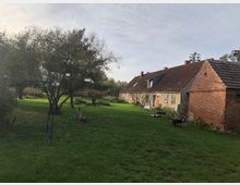 Großer ländlicher Garten mit einem alten, länglichen Gebäude aus roten Ziegeln und verputzten Wänden im Hintergrund. Im Vordergrund befinden sich Bäume, eine Rasenfläche und ein blaues Fußballtor.