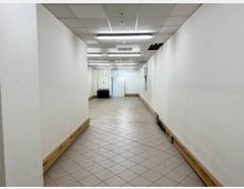 Ein leerer, schmaler Raum mit weißen Wänden, einer gefliesten Bodenfläche und einem abgehängten Deckenraster mit Leuchtstofflampen. Die Wände zeigen Gebrauchsspuren, und im Hintergrund sind ein weißer Schrank sowie ein schwarzes Möbelstück sichtbar.