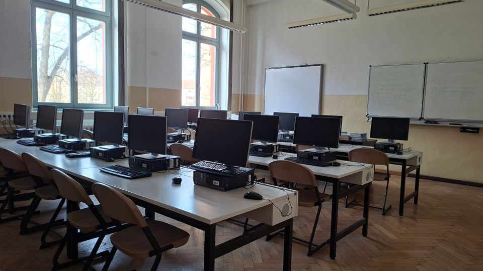 Computerraum/Klassenzimmer mit mehreren Reihen von Tischen, darauf Monitore, Tastaturen und Desktop-PCs, dazu Stühle. Große hohe Fenster lassen Tageslicht hinein; vorne an der Wand hängen Whiteboard und Tafel unter Deckenleuchten.