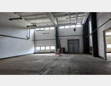 Leerer, industrieller Innenraum mit hohen Decken, weißen Wänden und Betonboden. Sichtbar sind Metallrohre, Oberlichter und Doppeltüren an einer der Wände.