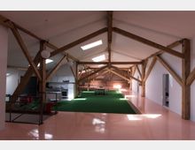 Geräumiger Dachbodenraum mit sichtbaren Holzbalken, weißen Wänden und einem glänzenden rosa Boden. In der Mitte des Raums befindet sich eine grüne Teppichfläche mit einem Tisch und Stühlen; im Hintergrund eine kleine Küchenzeile und eine Backsteinwand.