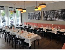 Ein gemütlich eingerichtetes Restaurant mit gedeckten Tischen, weißen Tischdecken und schwarzen Stühlen. Große Fenster mit weißen Vorhängen lassen Tageslicht herein, an der Wand hängen moderne, schwarz-weiße Kunstwerke, und die Decke ist mit mehreren Hängelampen beleuchtet.