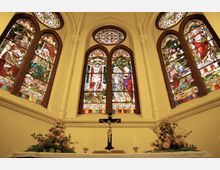 Innenraum einer Kirche mit einem Altar im Vordergrund, geschmückt mit Blumenarrangements. Im Hintergrund befinden sich große Spitzbogenfenster mit farbigem Glas, die biblische Szenen darstellen, sowie ein zentrales Kruzifix vor den Fenstern.