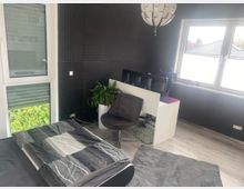 Modernes Schlafzimmer mit dunkler, strukturierter Wandverkleidung, einem Bett mit grauer Bettwäsche und hellem Laminatboden. Daneben stehen ein weißer Schreibtisch mit Monitoren, ein grauer Stuhl, eine Zimmerpflanze sowie große Fenster und eine Glastür mit Rollläden.