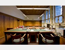 Ein Konferenzraum mit einer Holztäfelung an den Wänden und großen Fenstern mit Gitterverglasung auf der rechten Seite. Der Raum enthält einen großen, grünen Tisch mit Bürostühlen, Computermonitoren und Büromaterialien unter einer Holzdecke mit hängenden Leuchtstofflampen.