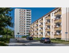 Wohngebiet mit einem hohen, mehrstöckigen Wohngebäude im Hintergrund und einem länglichen weißen Wohnhaus mit gelben Balkonen im Vordergrund. Autos sind entlang einer asphaltierten Straße geparkt, und Bäume sowie Grünflächen säumen den Bereich.