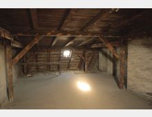 Ein Dachbodenraum mit sichtbaren Holzbalken und einer Holzdecke, einer kleinen Fensteröffnung, die Tageslicht hereinlässt, und einer Wand aus rauem Mauerwerk. Der Boden ist mit einfachen Holzdielen bedeckt, und der Raum wirkt unrenoviert.