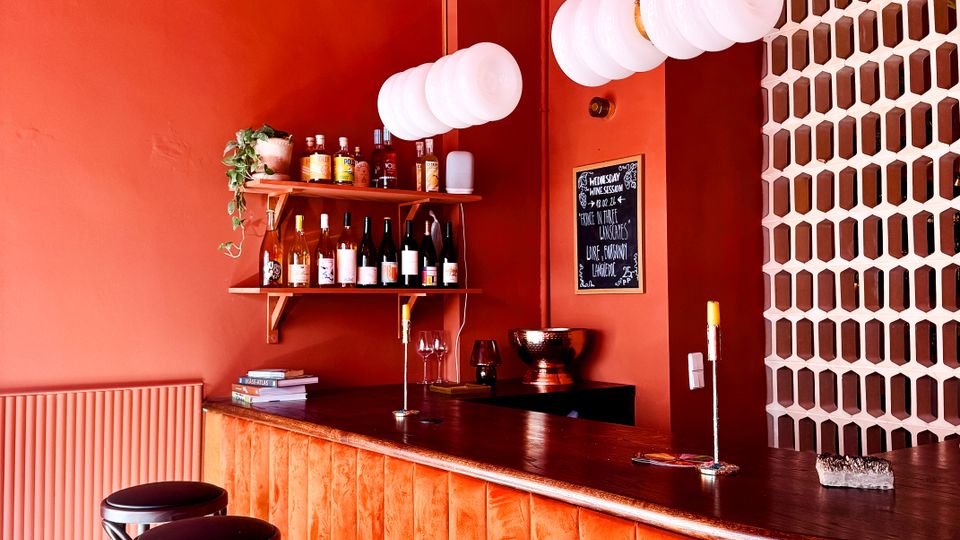 Small bar interior with red-orange walls, a wooden counter lined with black stools, and white globe pendant lights hanging overhead. Shelves behind the bar display bottles and glassware, with a geometric white partition wall on the right.