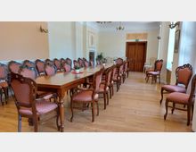 Raum mit langen Holztischen und geschnittenen Holzstühlen mit gepolsterten, gemusterten Rückenlehnen und Sitzflächen in rosa Farbton. Der Raum hat einen Parkettboden, helle Wände, verzierte Wanddetails, einige Pflanzen und eine Holztür am Ende.