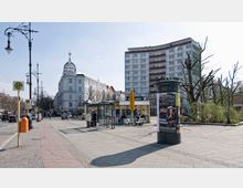 Straßenszene mit breitem Gehweg, Haltestellenzeichen für den öffentlichen Nahverkehr, einem Café mit gelber Markise und Menschen an Tischen. Im Hintergrund stehen ein historisches weißes Gebäude mit Kuppel und ein modernes Mehrfamilienhaus; kahle Bäume rahmen den Platz ein.