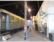 Innenraum eines historischen Bahndepots mit einem grünen Waggon auf Gleisen und einer weißen Wand mit einer analogen Uhr. Rechts führt eine Treppe zu einer erhöhten Plattform.