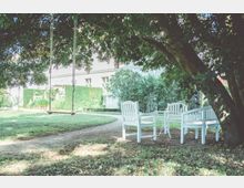 Ein Garten mit einer Holzschaukel, die an einem großen Baum hängt, und weißen Gartenmöbeln auf einer Grasfläche. Im Hintergrund ist ein Gebäude mit hellen Wänden und üppigen Hecken zu sehen.