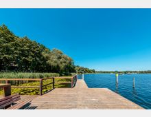 Holzsteg an einem ruhigen See mit Seerosen im Vordergrund, umgeben von Bäumen und Schilf am Ufer; Boote und Pfosten im Wasser sowie klarer blauer Himmel im Hintergrund.