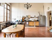 Eine großzügige, rustikale Küche mit einem hölzernen Esstisch und Stühlen im Vordergrund, einer Küchenzeile mit offenen Regalen und Holzschränken an der Rückwand sowie einem großen, silbernen Kühlschrank auf der rechten Seite. Ein Kronleuchter hängt von der Decke, und auf der linken Seite befinden sich große, geöffnete Fenster, die viel Tageslicht hereinlassen.