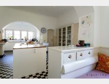 Helle Küche mit einem schwarz-weiß karierten Boden, einem großen weißen Küchenblock mit Holzarbeitsplatte in der Mitte, sowie weißen Schränken und Regalen an den Wänden. Ein großes Fenster mit vielen Pflanzen lässt Tageslicht herein, und an der Wand hängen Kinderzeichnungen.