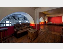 Innenraum eines Veranstaltungsraums mit Holzboden und roten Wänden. Auf der linken Seite steht ein braunes, gepolstertes Ledersofa vor einer halbrunden Fensterfront mit Bühnenscheinwerfern; im Hintergrund rechts ist eine kleine Bühne sichtbar.