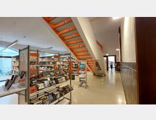 Ein moderner Innenraum einer Bibliothek mit Bücherregalen voller Bücher, einer markanten, freitragenden Treppe mit orangefarbenen Elementen und glatten cremefarbenen Böden. Im Hintergrund führt eine offene Tür zu einem weiteren Raum, und eine Person ist im Durchgang erkennbar.