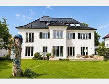 Großes weißes Wohnhaus mit dunklem Walmdach, Dachfenstern und Solarpaneelen, symmetrischer Fensterfront mit schwarzen Fensterläden und mehreren Terrassentüren. Davor liegt ein weitläufiger Rasen mit Terrasse und Gartenmöbeln sowie einem Kletterstamm am linken Bildrand.