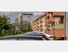 Wohngebiet mit mehrstöckigen Wohngebäuden, darunter ein graues modernes Gebäude mit Solarpaneelen auf dem Dach und orange-beigen Gebäuden mit Balkonen. Im Vordergrund geparkte Autos und vereinzelte Bäume.