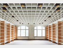 Ein leerer Raum mit Holzregalen an den Seitenwänden, hohen Fenstern mit Rundbögen und einer Decke mit einem gitterartigen Raster und eingebauten zylindrischen Leuchten. Der Boden ist mit Teppichboden bedeckt, und die Fenster lassen Tageslicht hinein.