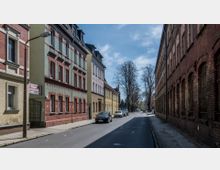 Straßenszene mit Altbauten: Links farbige Wohnhäuser mit rotem Backstein und Stuckdetails, rechts ein großes, schlichtes Backsteingebäude mit Rundbogenfenstern; geparkte Autos am Straßenrand sowie Bäume im Hintergrund.