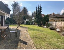 Gepflegter Garten mit Rasenfläche, Pflastersteinterrasse und Gartenmöbeln. Im Hintergrund sind Bäume, Sträucher, ein Gartenhaus sowie Nachbarhäuser zu sehen, umgeben von einem weißen Zaun.