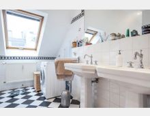 Badezimmer mit Dachfenster, weiß gefliesten Wänden und schwarzer Bordüre. Der Raum enthält zwei Waschtische, eine Waschmaschine, einen Heizkörper und einen schwarz-weiß karierten Boden.