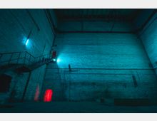 Innenansicht einer großen, leeren Industriehalle mit hohen Ziegelwänden und sichtbaren Stahlträgern an der Decke. Eine beleuchtete Metalltreppe führt seitlich zu einem roten beleuchteten Durchgang, während das Blau von Neonleuchten die Wände in kühles Licht taucht.