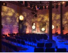 Ein Veranstaltungsraum in einem rustikalen Gebäude mit freiliegenden Steinwänden, mehreren beleuchteten, runden Lampen, die an den Wänden befestigt sind, und einer Bühne im Hintergrund. Im Vordergrund sind mehrere Reihen mit leeren Stühlen zu sehen, und die Beleuchtung erzeugt farbige Akzente in Blau und Orange.