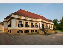 Historisches Herrenhaus mit gelb-weißer Fassade, roten Ziegeldach und symmetrischen schwarzen Fensterläden. Eine geschwungene Steintreppe führt zu einem Haupteingang mit Rundbogen, umgeben von Bäumen und einer Kiesfläche.