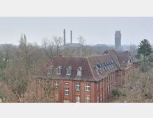 Backsteingebäude mit steilem Satteldach und mehreren Dachgauben, umgeben von kahlen Bäumen in einer winterlichen Landschaft. Im Hintergrund sind zwei Industrieschornsteine und ein hoher Wasserturm zu sehen.