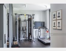 Moderner Fitnessraum mit einem Multifunktionsgerät in der Mitte, einem Laufband rechts und einem Flachbildfernseher an der Wand. Der Raum hat helle Wände, dunklen Holzboden, gerahmte Schwarz-Weiß-Bilder an der rechten Wand und große Fenster, die natürliches Licht hereinlassen.