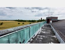 Ansicht eines Flachdachs mit verwitterter grüner Brüstung und technischen Installationen, darunter eine Satellitenschüssel und Kabel. Im Hintergrund ist eine weite, baumbestandene Feldlandschaft unter bewölktem Himmel zu sehen.