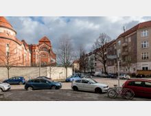 Straßenszene in einem Wohnviertel mit mehrstöckigen Wohnhäusern auf der rechten Seite und einem markanten roten Ziegelgebäude mit Türmen und Verzierungen auf der linken Seite. Im Vordergrund stehen geparkte Autos und Fahrräder an einer gepflasterten Straße, während kahle Bäume die Szenerie ergänzen.