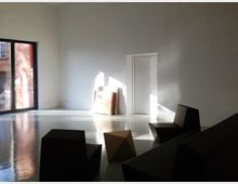 Minimalistisch eingerichteter Raum mit weißen Wänden, großem Fenster auf der linken Seite, das Tageslicht hereinlässt, und dunklen geometrischen Möbelstücken. Im Hintergrund steht eine Tür, daneben sind einige Flachgegenstände an die Wand gelehnt.