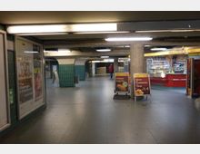 Unterirdische Fußgängerpassage mit grünen Fliesen an den Wänden, Werbeplakaten und Verkaufsständen, darunter eine Bäckerei mit leuchtend roten Theken und Aufstellern für Angebote. Die Passage ist schwach beleuchtet, mit Betonpfeilern und gewölbten Durchgängen.
