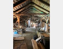 Dachboden in einem rustikalen Gebäude mit Holzbalken und Holzdielen, genutzt als Werkstatt oder Lagerraum. Verschiedene Baumaterialien, Werkzeuge und Möbel sind unordentlich verteilt, mit natürlichem Licht, das durch kleine Fenster einfällt.