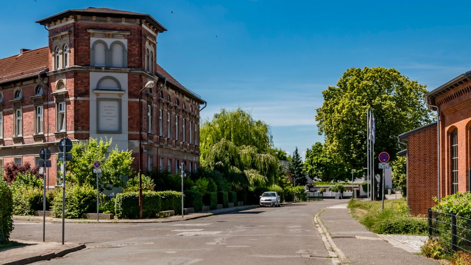 Straßenecke mit einem mehrstöckigen historischen Backsteingebäude mit Turmaufsatz und Rundbogenfenstern, umgeben von Hecken und Bäumen. Rechts steht ein weiteres Backsteingebäude entlang einer ruhigen Straße unter blauem Himmel.