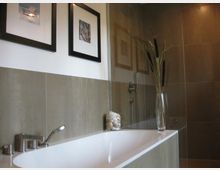 Modernes Badezimmer mit einer weißen Badewanne, umgeben von beige-grauen Fliesen. An der Wand hängen zwei gerahmte Kunstbilder, daneben steht eine Glasvase mit dekorativen Zweigen.