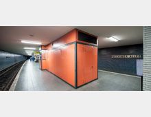 U-Bahn-Station mit orangefarbenem Technikraum in der Mitte des Bahnsteigs, blauen Wandfliesen und einem Schild mit der Aufschrift „Parchimer Allee“ an der rechten Wand. Links verläuft das Gleis neben einem gewölbten Bahnsteig.