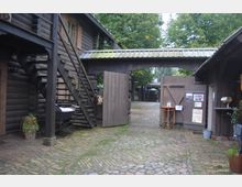 Ein rustikaler Hof mit Holzhäusern und offenen Holztoren, die auf einen gepflasterten Innenhof führen. Eine Holztreppe führt zu einem oberen Bereich, und umgeben ist die Szene von Bäumen und dekorativen Elementen.