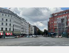 Straßenszene in einer städtischen Umgebung mit mehrstöckigen Gebäuden im Hintergrund, darunter ein weißes Hotelgebäude und ein rotes Backsteingebäude, teils mit Baugerüsten bedeckt. Im Vordergrund sind Straßenbahnschienen, Ampeln, parkende Autos und Gehwege sowie ein Eingang zur U-Bahn („U“-Schild) sichtbar.