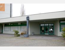 Flacher, moderner Gebäudeeingang mit überdachtem Vorbereich, grünen Fensterrahmen und einer verglasten Eingangstür, umgeben von Pflanzkübeln und einem gepflasterten Vorplatz.