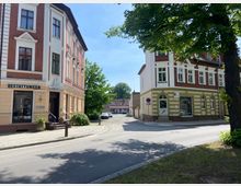 Straßenszene in einer Kleinstadt mit zwei mehrstöckigen Gebäuden aus Backstein und Putzfassaden; links ein Bestattungsunternehmen, rechts ein Eckgebäude mit Schaufenstern. Im Hintergrund eine schmale Gasse mit geparkten Autos und kleinen Häusern.