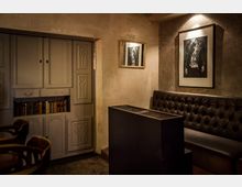 Gemütlicher Innenraum mit dunkler Ledersitzbank an einer rauen, verputzten Wand. Zwei gerahmte Schwarz-Weiß-Bilder hängen an der Wand, und ein eingebautes Regal mit Büchern ist in eine Schrankwand integriert.