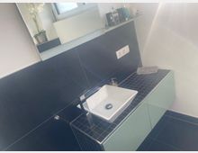 Modernes Badezimmer-Waschbecken auf einer dunkel gefliesten Ablage mit mintfarbenen Unterschränken und schwarzer Wandverkleidung. Darüber befindet sich ein Fenster mit kleiner Ablagefläche, auf der verschiedene Pflegeartikel stehen.