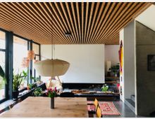 Ein modernes Wohnzimmer mit offener Gestaltung und großen Glasfenstern, die viel Licht hereinlassen. Sichtbare Elemente sind eine Holzlatten-Decke, ein hölzerner Esstisch mit Blumen, ein hängender Lampenschirm, zahlreiche Pflanzen sowie eine minimalistische Sitzecke mit weißen Wänden und farbigen Akzenten.