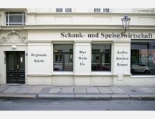 Fassade eines traditionellen Restaurants mit der Aufschrift „Schank- und Speisewirtschaft“ auf cremefarbenem Putz. Das Gebäude hat dekorative Elemente über der Eingangstür, drei Fenster mit Schriftzügen wie „Regionale Küche“, „Bier, Wein, Eis“ und „Kaffee, Kuchen, Brause“ sowie eine historische Laterne an der Wand.